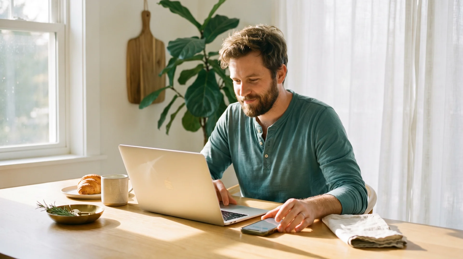 Freelancer at a sunlit kitchen table, laptop open, phone face-up next to him — moving fluidly between clicking and asking